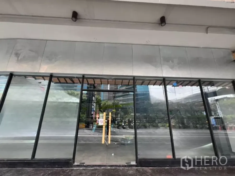 พื้นที่ขายปลีก ให้เช่า - หน้าร้านกระจกเต็มบานระดับทางเท้า พร้อมประตูคู่ ใต้กันสาด สะท้อนอาคารโดยรอบ.