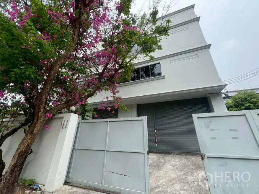 Warehouse for rent in Phra Khanong, Bangkok - Front elevation of warehouse with wide steel gates and roller shutter under pink blossoms