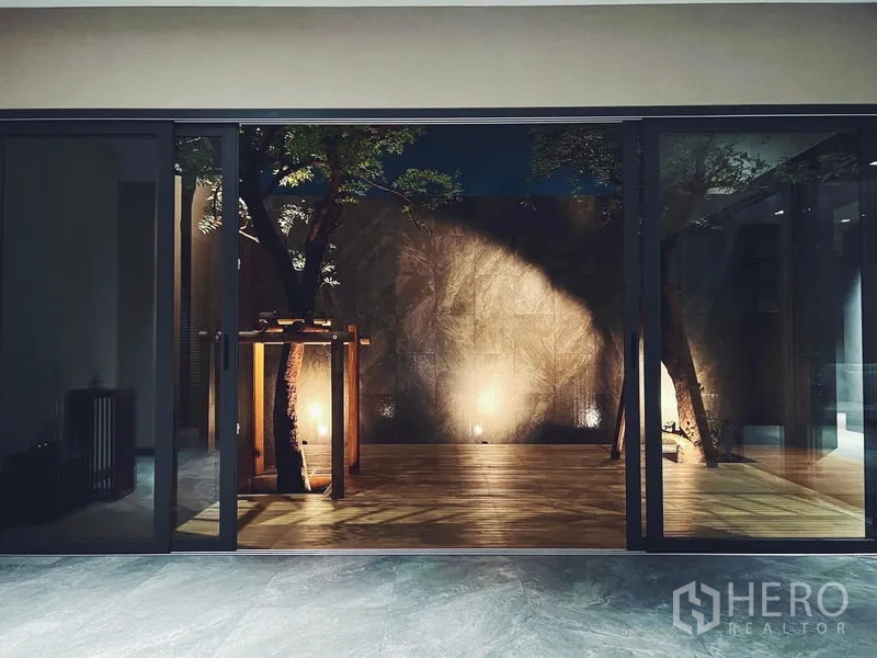 บ้าน ให้เช่าหรือขายในBangkok - เฉลียงคอร์ตยาร์ดยามค่ำ พร้อมต้นไม้และไฟส่องสว่าง มองผ่านประตูกระจก