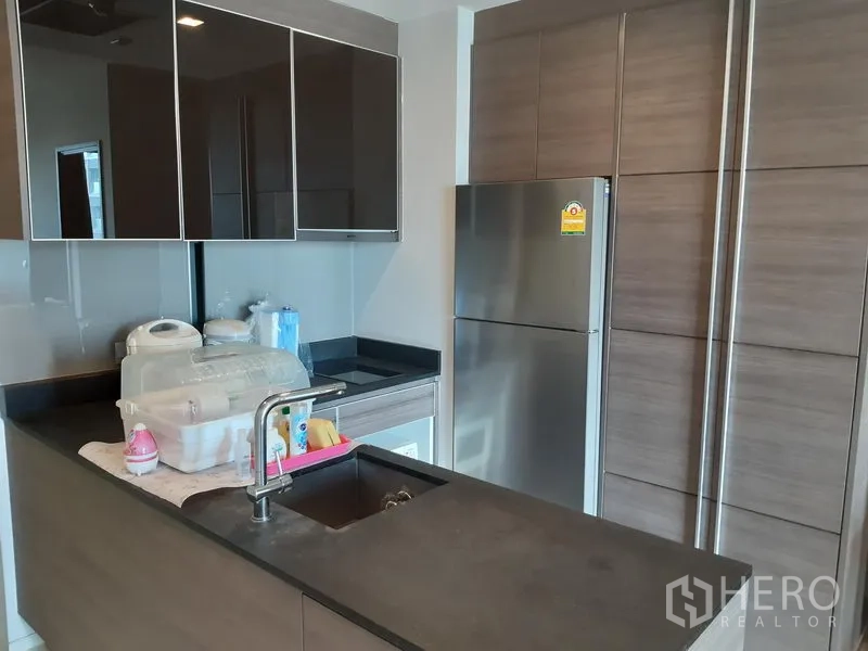 公寓 出租在Khlong Toei, Bangkok - 现代小厨房，深色台面、亮面吊柜与不锈钢冰箱。
