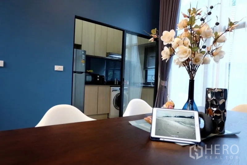Townhouse for rent in Khlong Toei, Bangkok - Dining table detail with flower vase, looking toward a glass-partitioned kitchen with light wood cabinets and a washer.