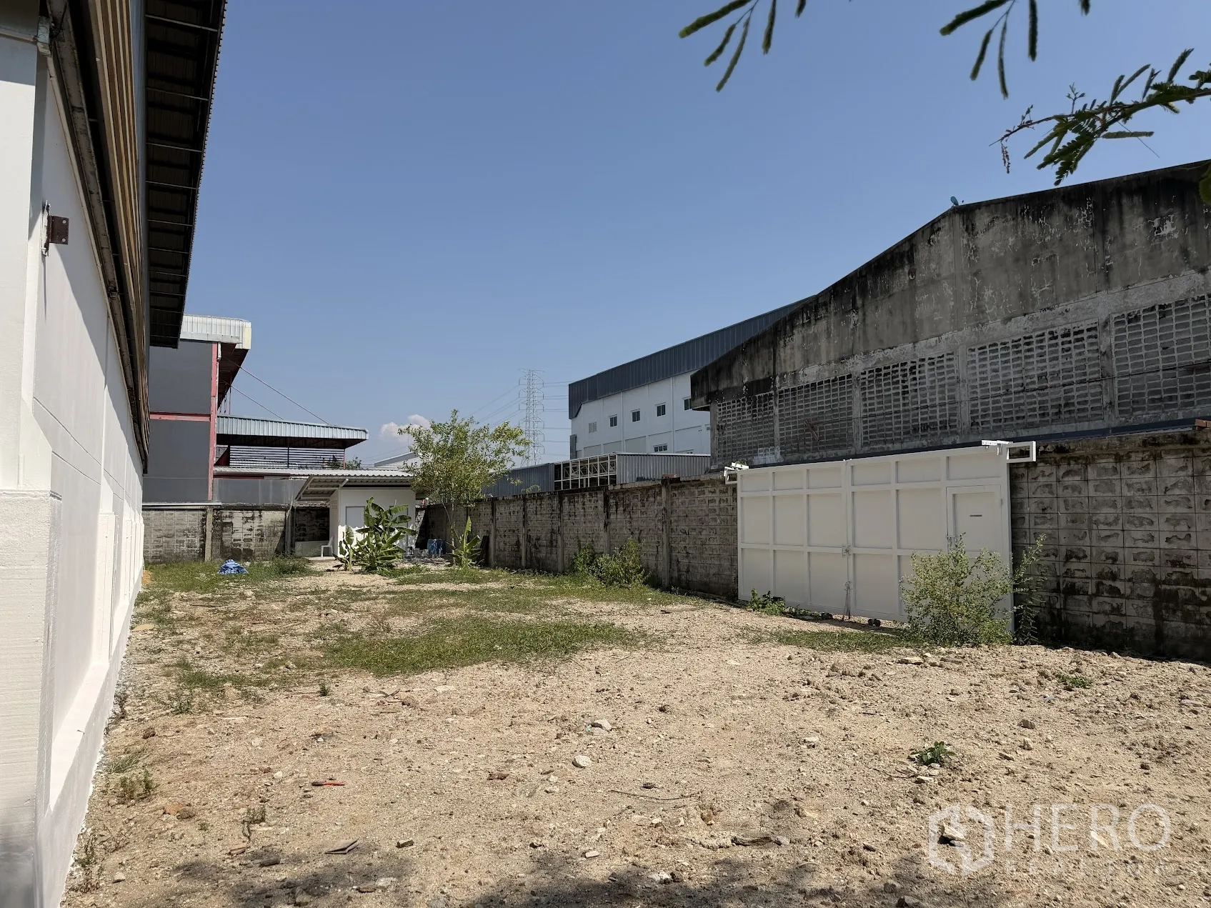 Factory for rent in Bang Sao Thong, Samut Prakan - Gated side yard with dirt surface, perimeter wall, and a small utility hut beside factory buildings.