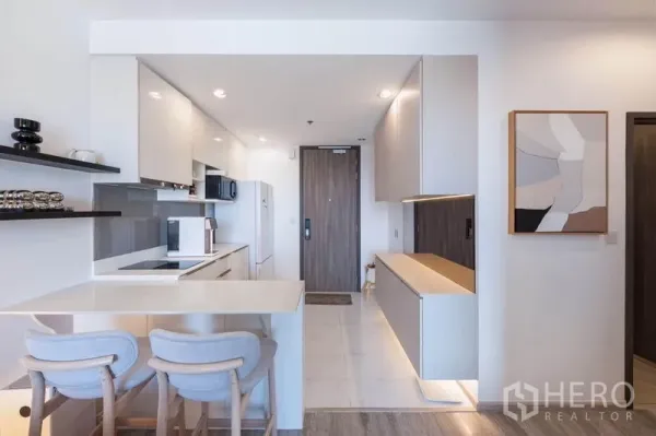 Condominium for rent in Bang Na, Bangkok - Modern white kitchen with breakfast bar, two stools, built-in cabinets, and the entry door.