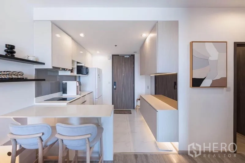 Condominium for rent in Bang Na, Bangkok - Modern white kitchen with breakfast bar, two stools, built-in cabinets, and the entry door.