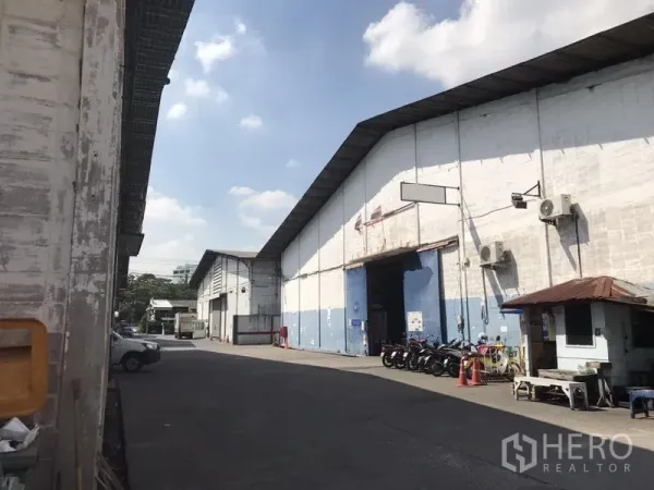 货栈 出租在Khlong Toei, Bangkok - 仓库外立面，超高卷帘门、带顶通道、停放的摩托车与门岗