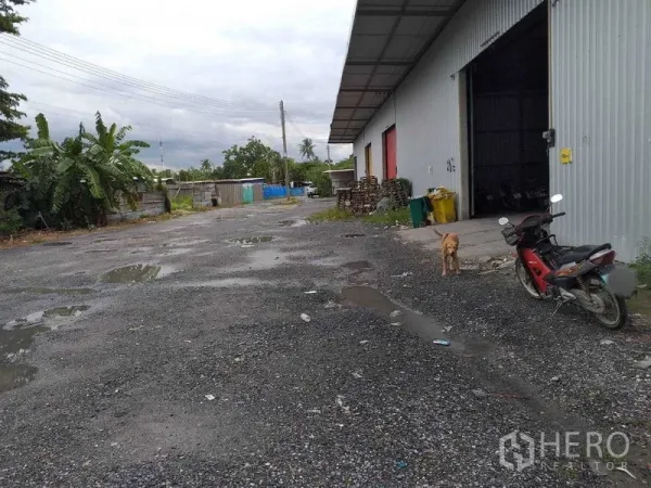 Warehouse for rent in Pak Kret, Nonthaburi - Gravel yard beside a metal-clad warehouse with long eaves and an open roller door in Pak Kret.