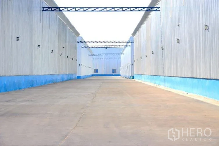 Warehouse for rent in Lat Lum Kaeo, Pathum Thani - Wide concrete driveway between metal-clad warehouse blocks with overhead trusses and lighting.
