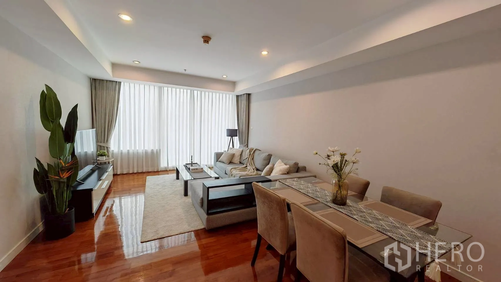 Condominium for rent in Khlong Toei, Bangkok - Open-plan living and dining room with sofa, TV and dining set by large sheer curtains.