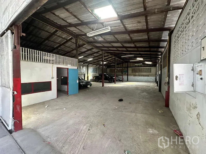 Warehouse for rent in Suan Luang, Bangkok - View from the entrance across a long-span warehouse with a small office unit and vehicles inside.
