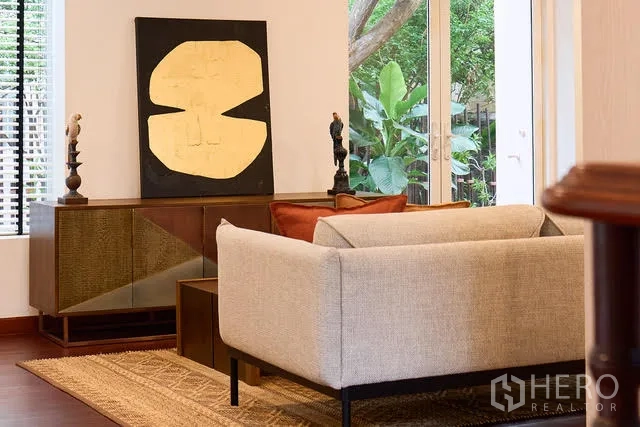 บ้าน ให้เช่าในWatthana, Bangkok - มุมนั่งเล่นอบอุ่น โซฟาสีอ่อน ตู้วางของ และงานศิลป์ใกล้ประตูกระจกเปิดสวน