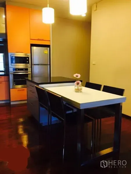 Condominium for rent in Watthana, Bangkok - Kitchen island with bar chairs, orange cabinetry, built-in oven and refrigerator under warm lighting.