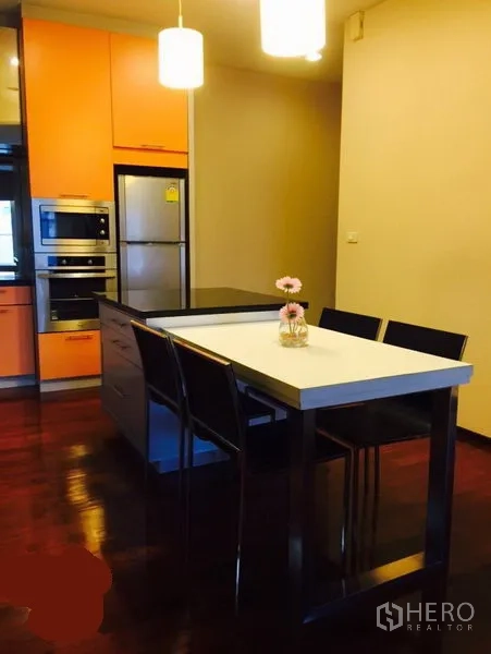 Condominium for rent in Watthana, Bangkok - Kitchen island with bar chairs, orange cabinetry, built-in oven and refrigerator under warm lighting.