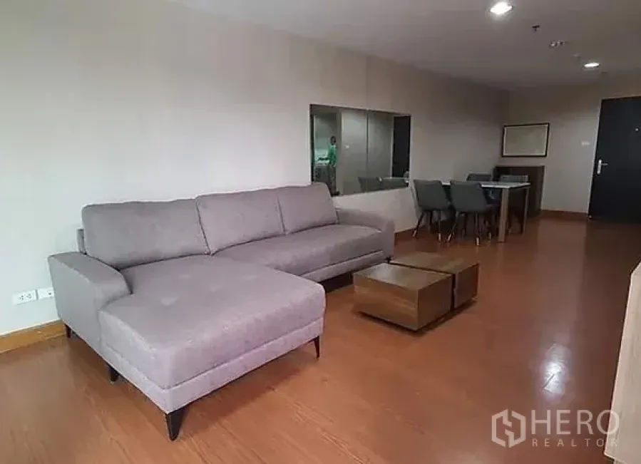 Condominium for rent in Huai Khwang, Bangkok - Spacious living room with gray L-shaped sofa, wood floors, coffee tables and a dining area.
