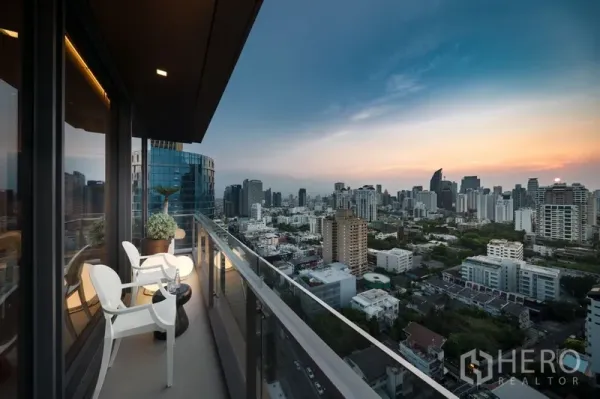 Condominium for sale in Watthana, Bangkok - High‑floor balcony with seating and glass railing overlooking city views at sunset.