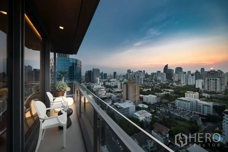 Condominium for sale in Watthana, Bangkok - High‑floor balcony with seating and glass railing overlooking city views at sunset.