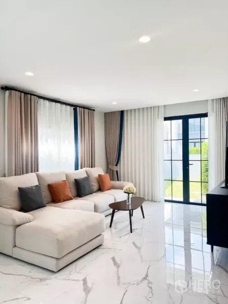 บ้าน ให้เช่าในKhan Na Yao, Bangkok - ห้องนั่งเล่นสว่างด้วยโซฟาเบจตัวใหญ่ พื้นกระเบื้องลายหินอ่อน และประตูกระจกเปิดสู่สวน