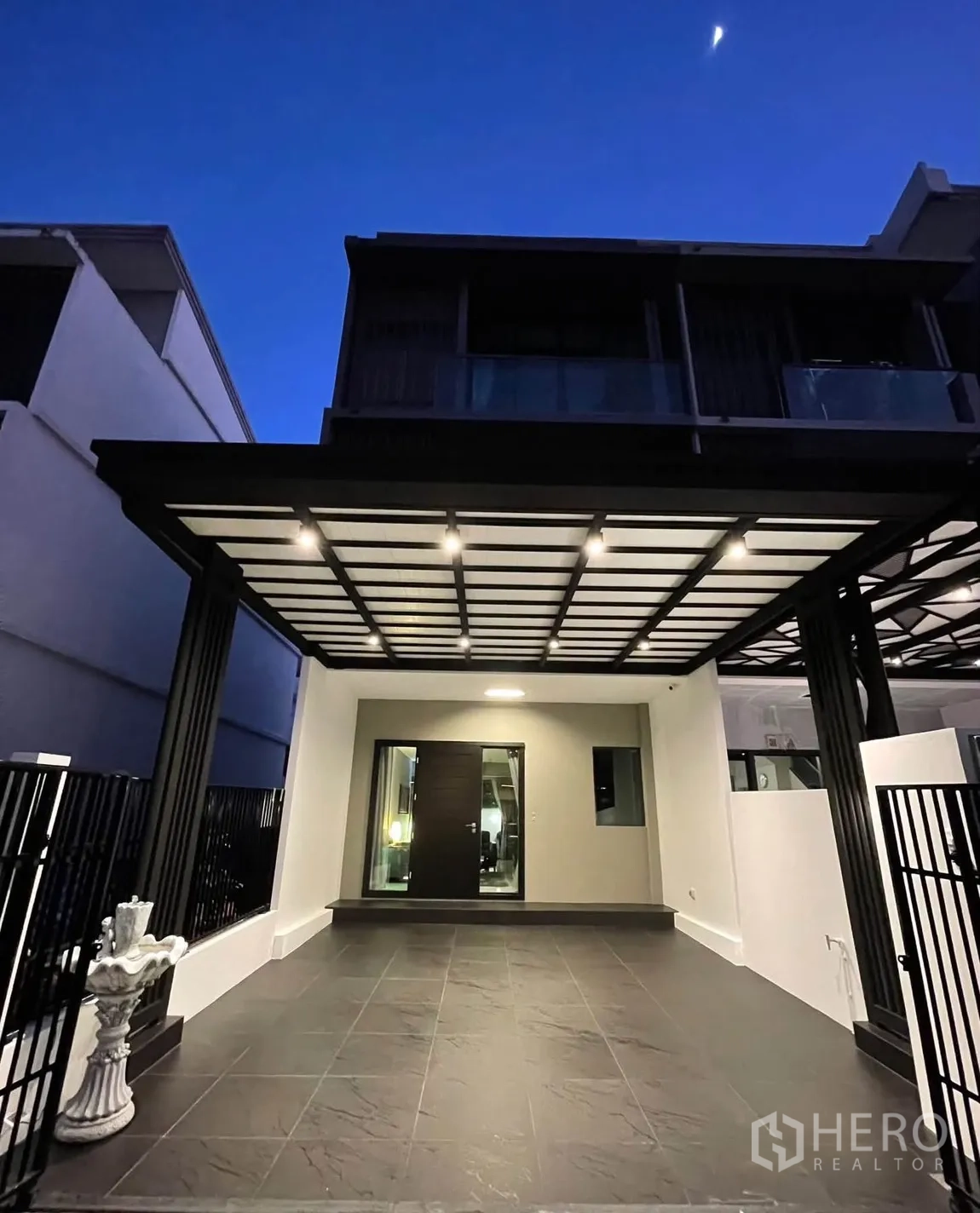 Townhouse for rent in Lat Phrao, Bangkok - Front carport with ceiling lights and gate at dusk.