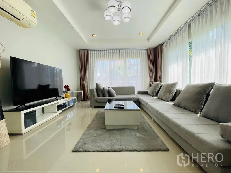 บ้าน ขายในLam Luk Ka, Pathum Thani - ห้องพักผ่อนกว้าง โซฟาเข้ามุมยาว ทีวีจอใหญ่ และหน้าต่างเต็มผนัง