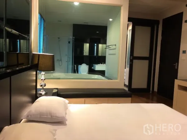 คอนโด ให้เช่าในPathum Wan, Bangkok - ห้องนอนหลักมีโคมไฟข้างเตียง และหน้าต่างกระจกมองเข้าสู่ห้องน้ำในตัวที่มีอ่างอาบน้ำและฝักบัว