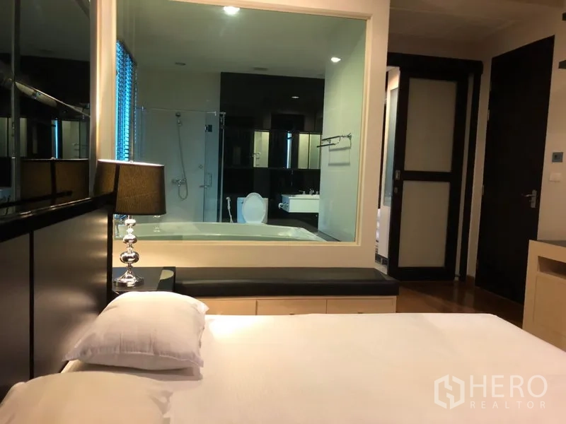 คอนโด ให้เช่าในPathum Wan, Bangkok - ห้องนอนหลักมีโคมไฟข้างเตียง และหน้าต่างกระจกมองเข้าสู่ห้องน้ำในตัวที่มีอ่างอาบน้ำและฝักบัว