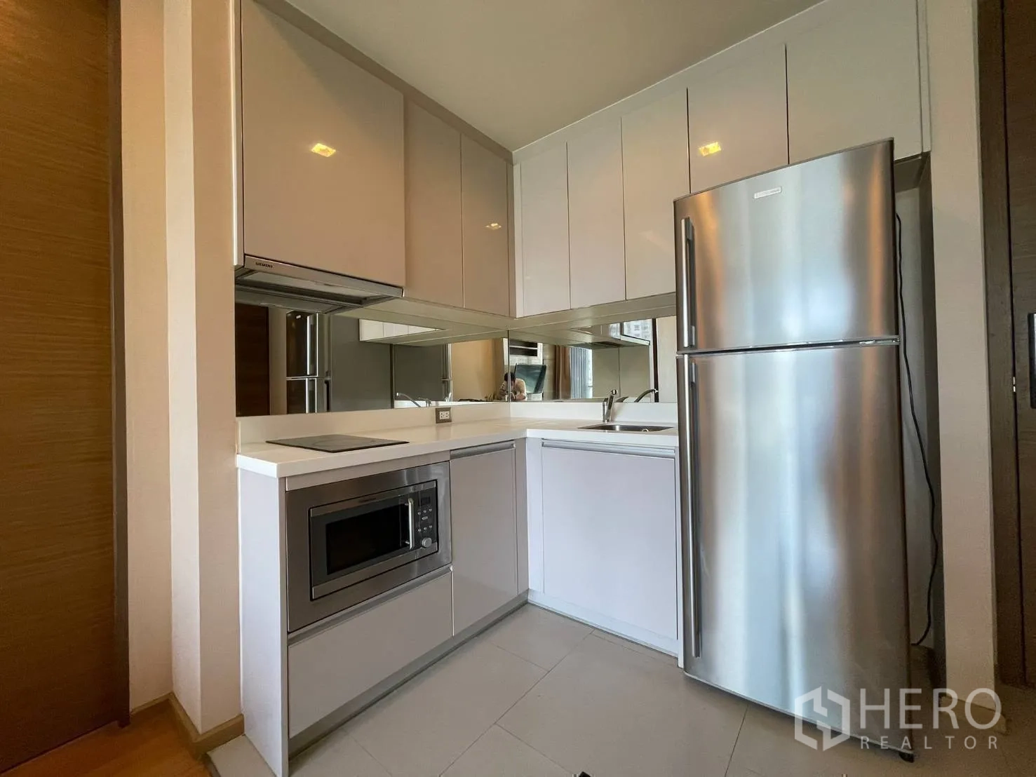 Condominium for rent or sale in Bang Rak, Bangkok - L-shaped kitchen with glossy cabinets, cooktop, microwave and stainless refrigerator.
