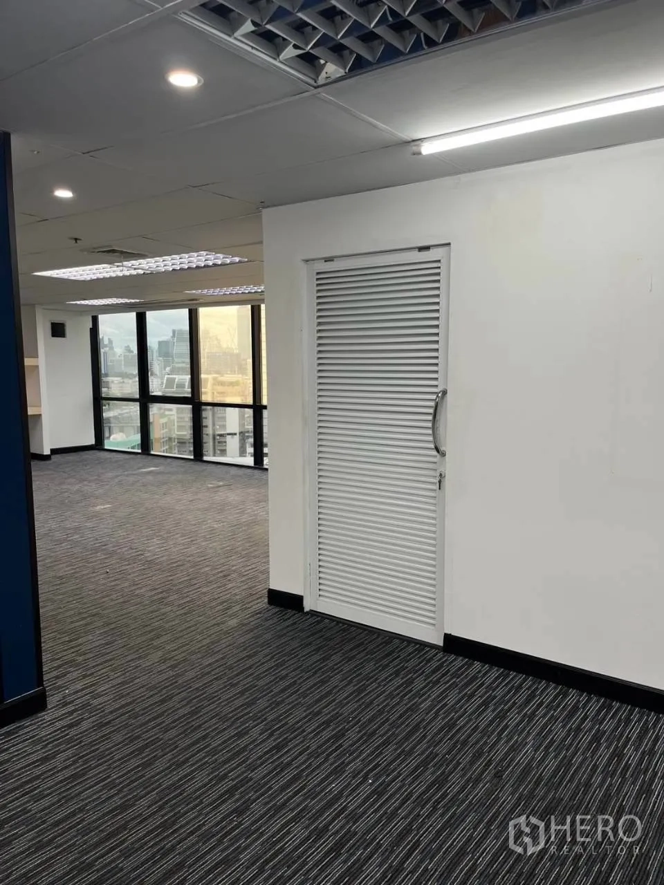 Office Space for rent in Khlong Toei, Bangkok - Open workspace with dark carpet, white louvered door, and distant city-view windows.