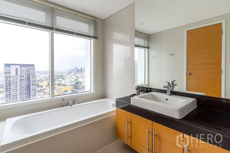 Condominium for rent in Watthana, Bangkok - Bathroom with soaking tub beside a large window and single-sink vanity, offering a high-floor city skyline view.