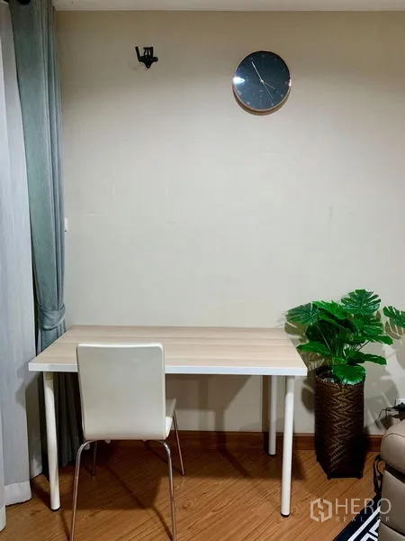Condominium for rent in Huai Khwang, Bangkok - Compact work corner with light wood desk, white chair, wall clock and potted plant.