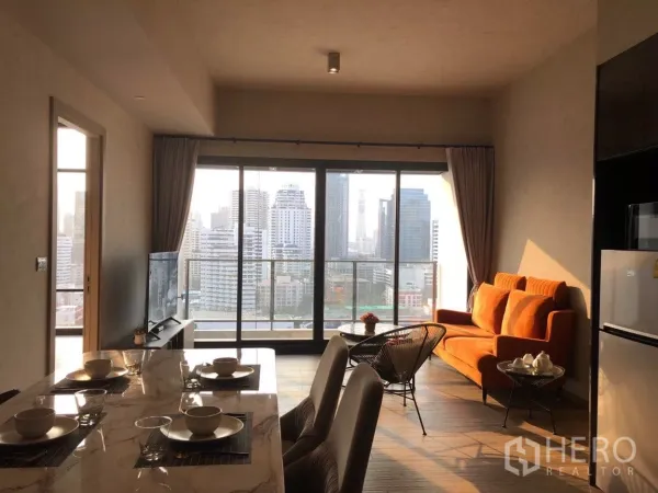 Condominium for sale in Watthana, Bangkok - Sunlit living and dining area with marble table, orange sofa, and floor-to-ceiling windows opening to a balcony with Bangkok skyline views.