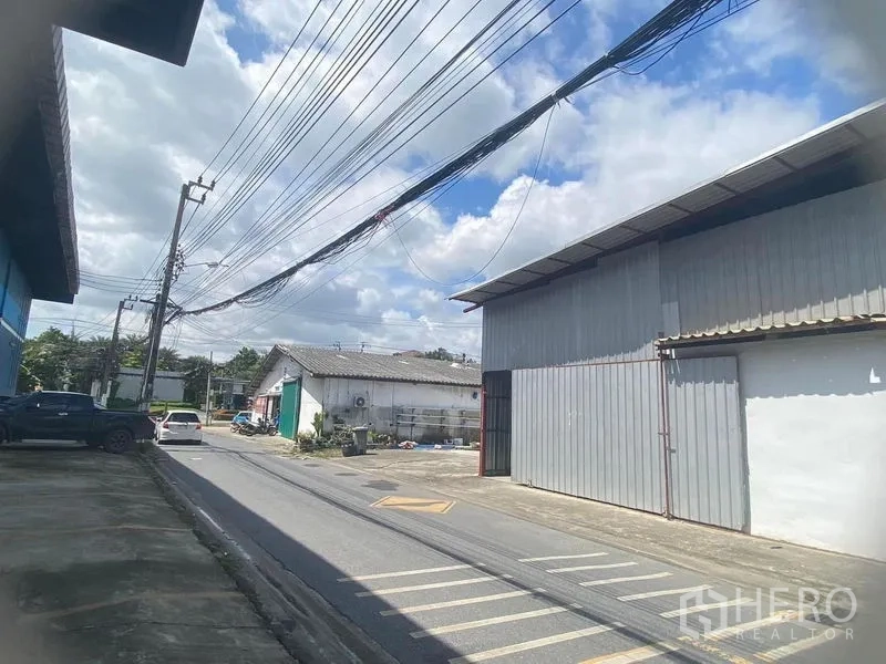 Warehouse for rent in Saphan Sung, Bangkok - Industrial lane with metal-clad warehouse frontage and overhead power lines under a blue sky, showing easy street access.