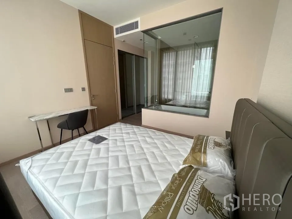 คอนโด ให้เช่า - ห้องนอนโปร่งสบาย หัวเตียงบุนวม โต๊ะทำงาน ตู้เก็บของ และผนังกระจกมองเข้าห้องน้ำ