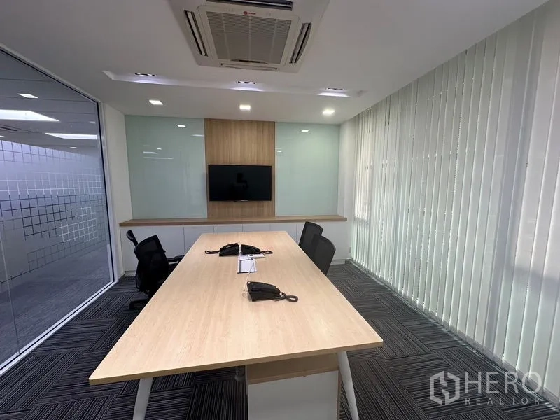 Office Space for rent - Meeting room with long table, wall-mounted TV, glass wall and floor-to-ceiling vertical blinds.