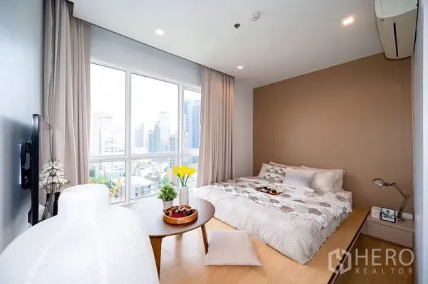 Condominium for rent or sale in Watthana, Bangkok - Corner bedroom with large windows, warm accent wall, and skyline views.