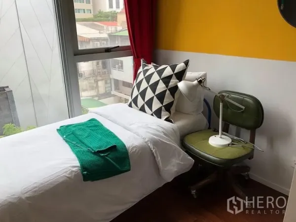Condominium for rent or sale in Watthana, Bangkok - Cozy single bedroom with large window, city view, colorful accent wall and a study chair with lamp.