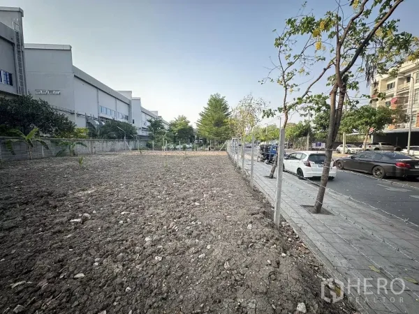 Land for sale in Saphan Sung, Bangkok - Wide street frontage of empty land with sidewalk, trees, and parked cars nearby.
