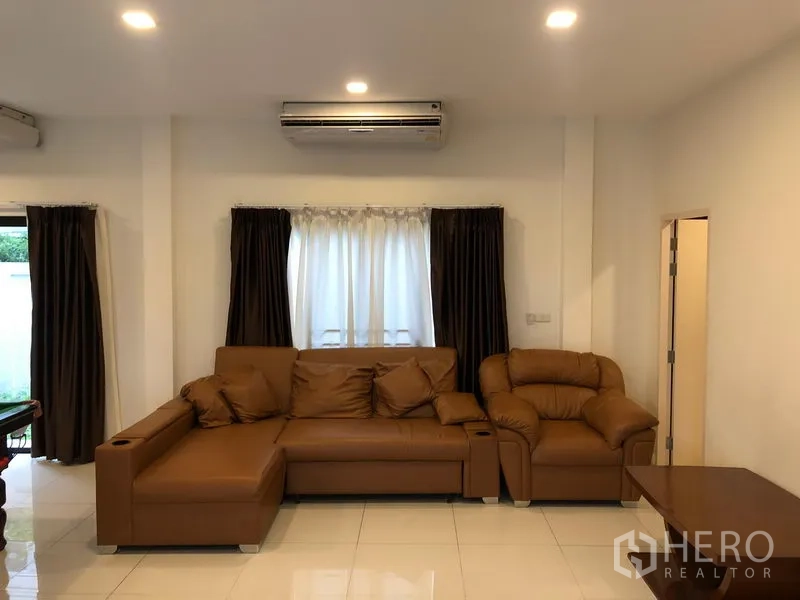 Townhouse for rent or sale in Saphan Sung, Bangkok - Cozy lounge with brown leather sectional sofa, window curtains and wall-mounted air conditioner.
