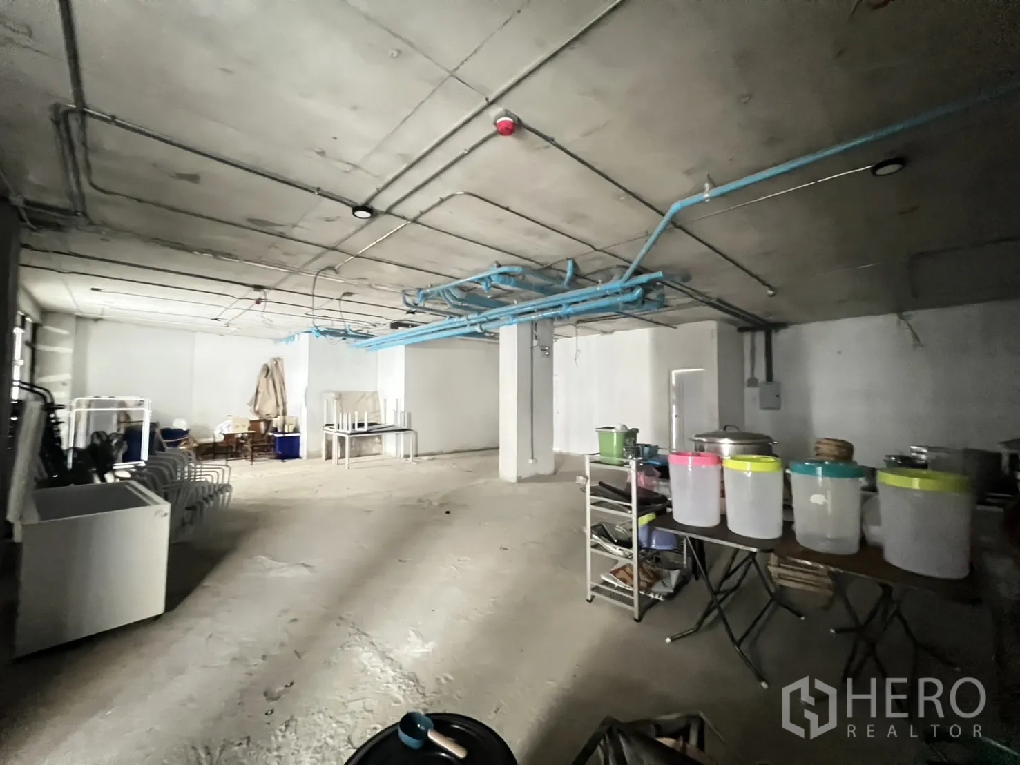 พื้นที่ขายปลีก ให้เช่า - พื้นร้านสภาพยังไม่ตกแต่ง มีอุปกรณ์เก็บของและเก้าอี้วางซ้อน ใกล้ประตูม้วนมีช่องแสง