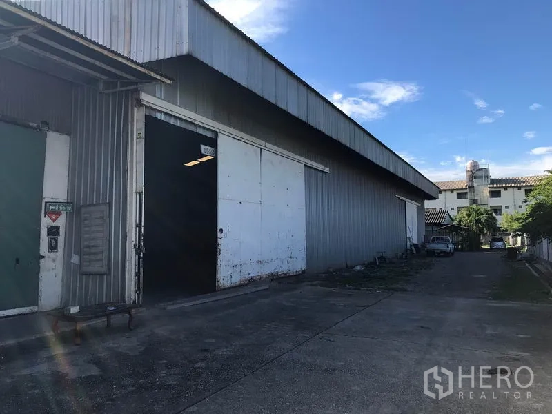 Warehouse for rent in Mueang Samut Sakhon, Samut Sakhon - Steel-clad warehouse exterior with large sliding door and driveway under blue sky.