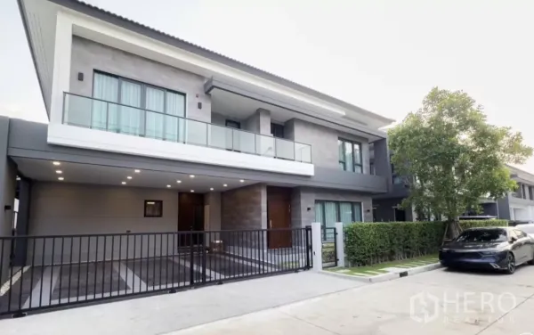 Detached House for rent in Bang Phli, Samut Prakan - Modern two-storey detached house with glass balcony, gated carport and tree-lined frontage.