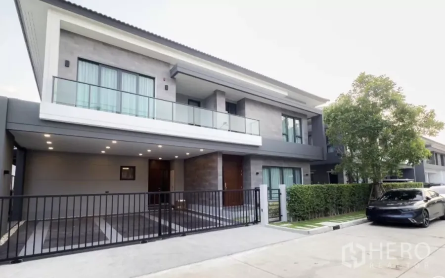 Detached House for rent in Bang Phli, Samut Prakan - Modern two-storey detached house with glass balcony, gated carport and tree-lined frontage.