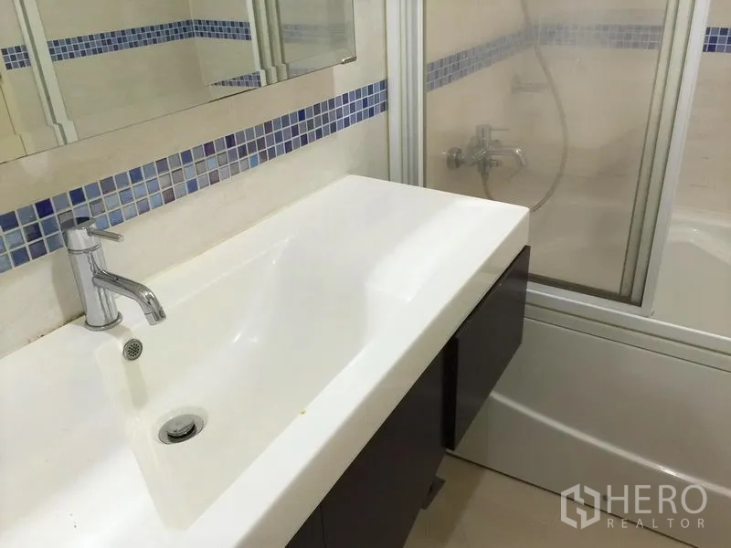 Condominium for rent in Watthana, Bangkok - Bathroom with long vanity, mirror, mosaic accent tiles and a bathtub with glass screen.