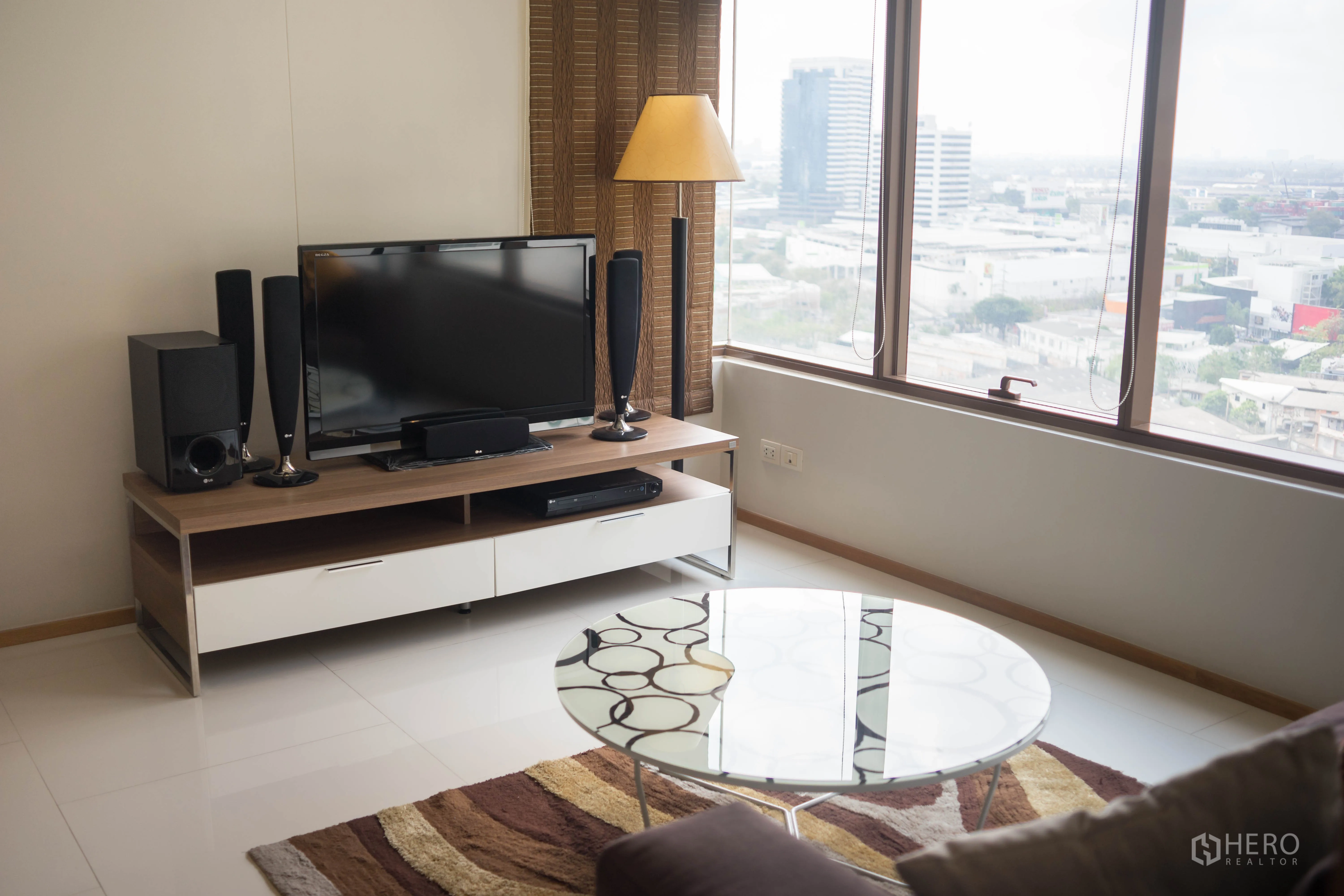 Condominium for rent or sale in Khlong Toei, Bangkok - Living room with TV console, floor lamp and wide windows showcasing city views.
