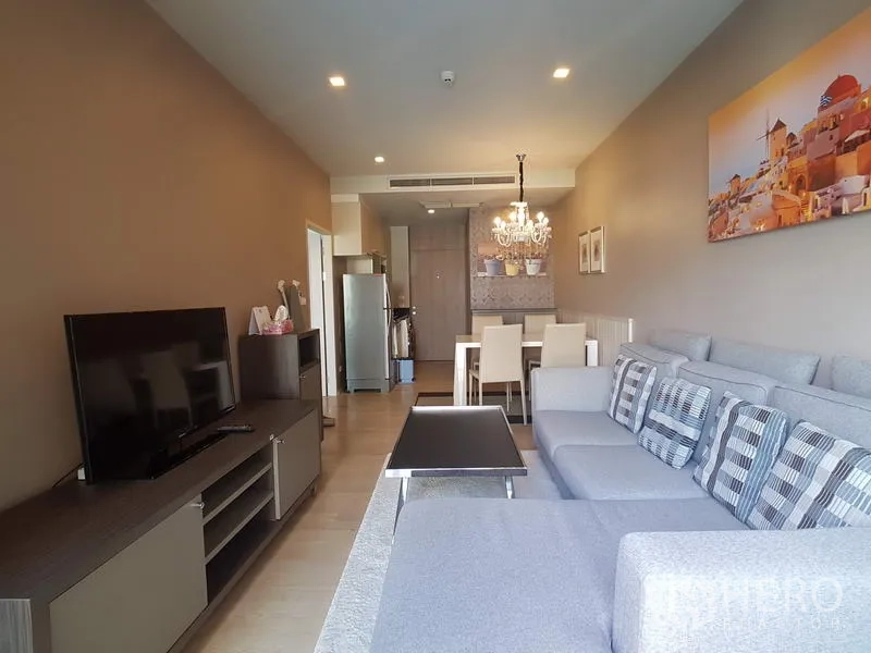 Condominium for rent in Bangkok - Open-plan living and dining area with L-shaped sofa, TV console and chandelier leading toward the kitchen.