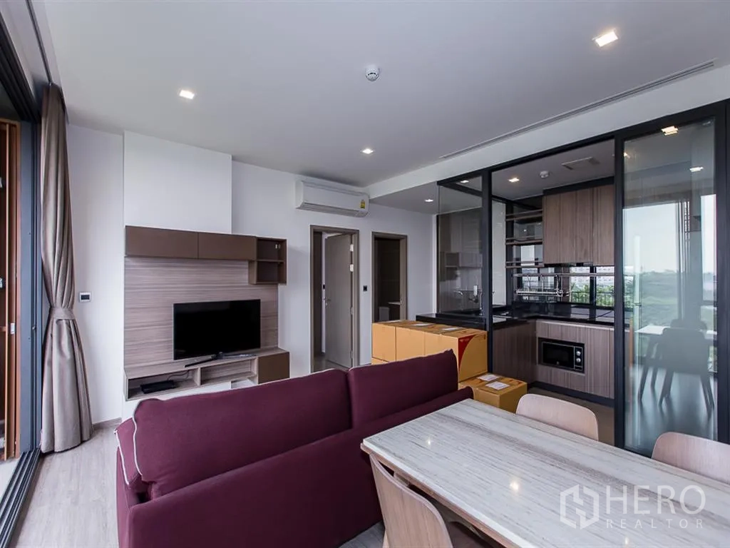 Condominium for rent in Watthana, Bangkok - Living room with TV console, maroon sofa, dining table, and glass-partitioned kitchen.