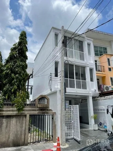 Townhouse for sale in Din Daeng, Bangkok - Side view of a modern white three-storey townhouse with balcony railings, driveway and opening gate.