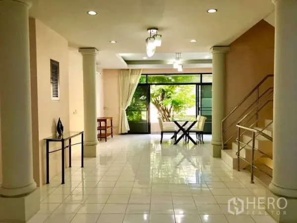 Townhouse for rent in Watthana, Bangkok - Open-plan living area with columns, staircase and sliding glass doors facing greenery.
