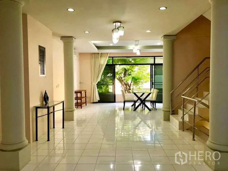 Townhouse for rent in Watthana, Bangkok - Open-plan living area with columns, staircase and sliding glass doors facing greenery.