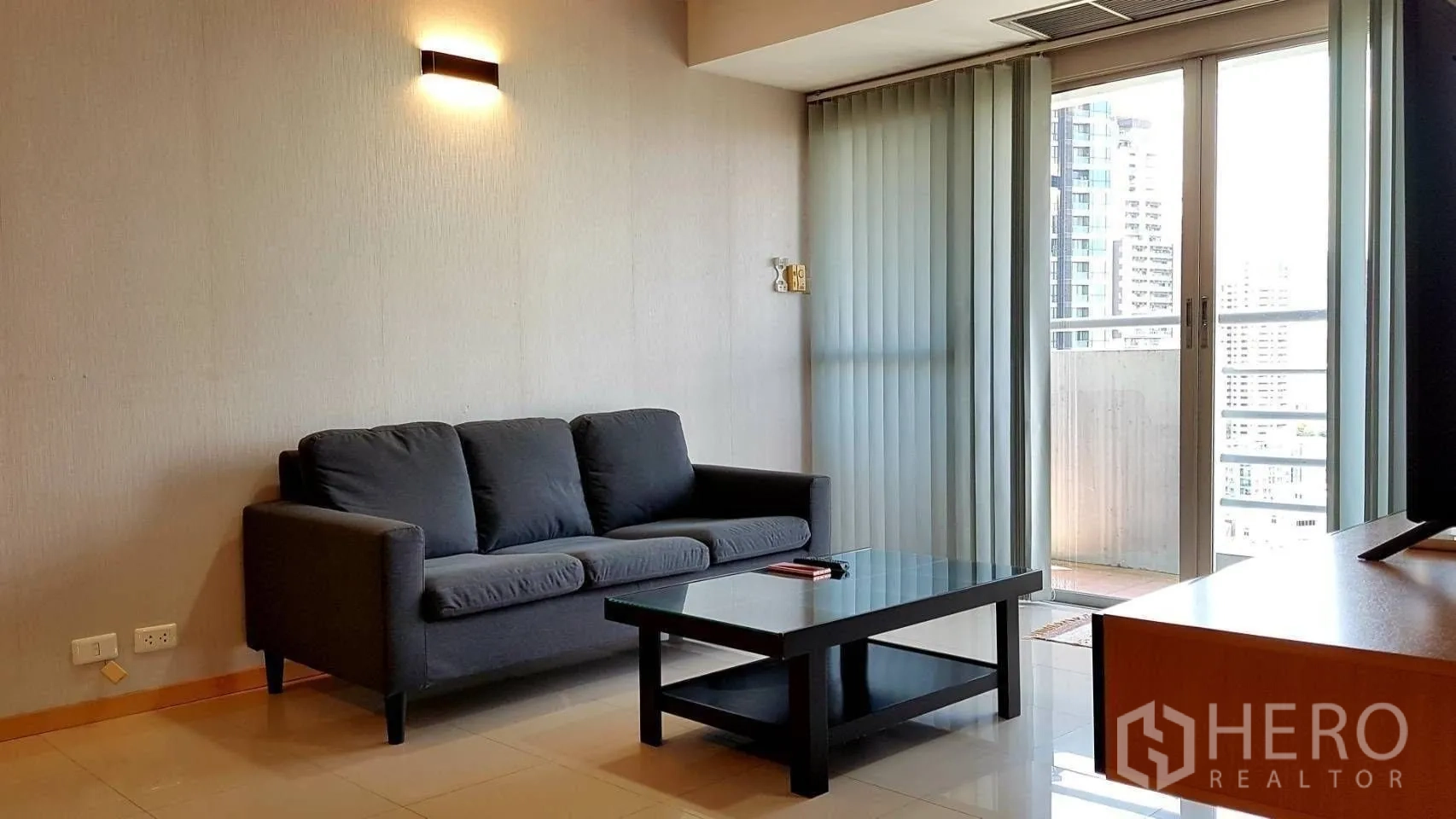 Condominium for rent in Khlong Toei, Bangkok - Bright living area with dark sofa, glass coffee table and balcony doors with city view.