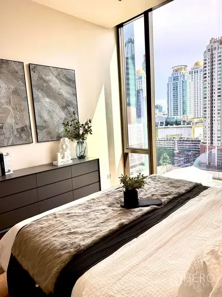 Condominium for rent in Pathum Wan, Bangkok - Bedroom showcasing a skyline view through full-height glass beside a dresser and wall art.