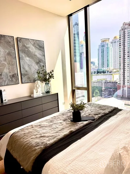 Condominium for rent in Pathum Wan, Bangkok - Bedroom showcasing a skyline view through full-height glass beside a dresser and wall art.